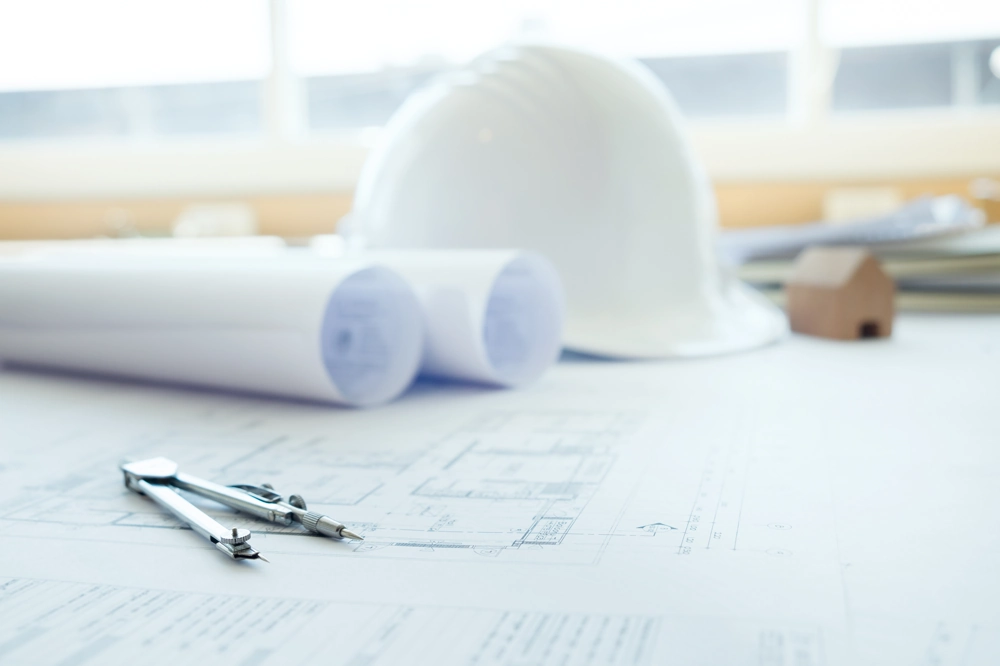 Projekt domu jednorodzinnego: rozłożone plany architektoniczne, cyrkiel i biały kask na stole